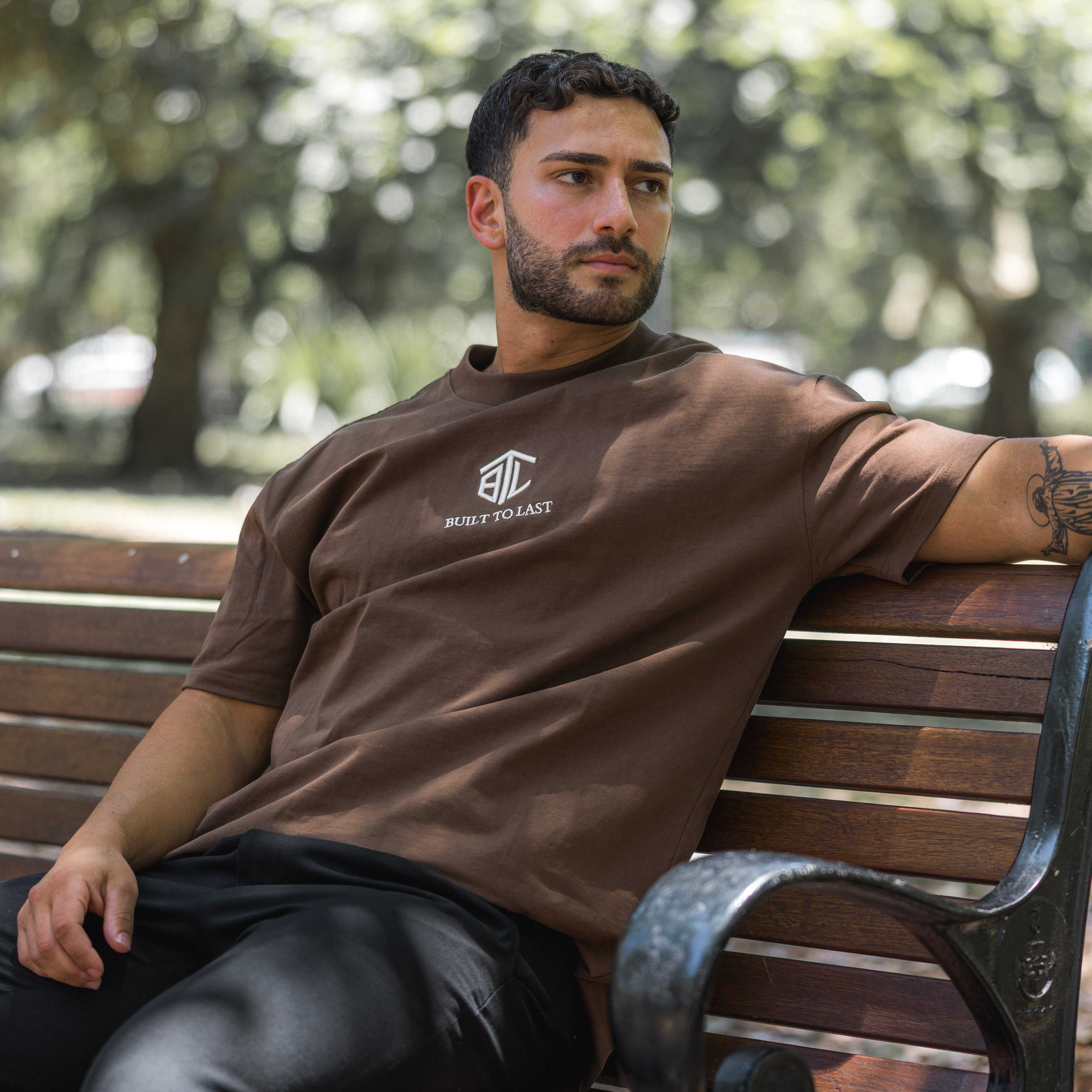 Model sitting on a wooden park bench wearing Built to Last Eternal Tee Chocolate heavyweight cotton T-shirt with embroidered chest logo