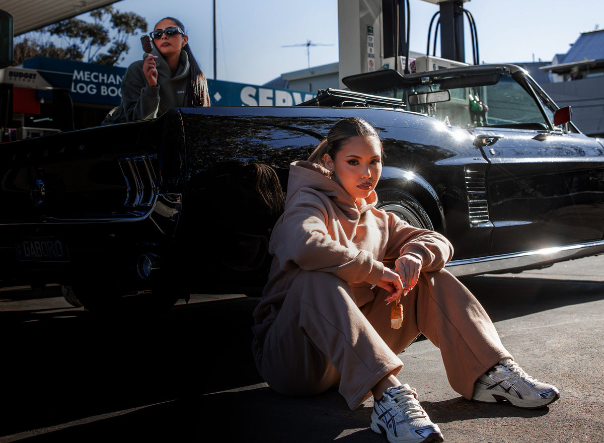 Women's sand oversized tracksuit set by Built To Last at a retro gas station. Classic black convertible Mustang.