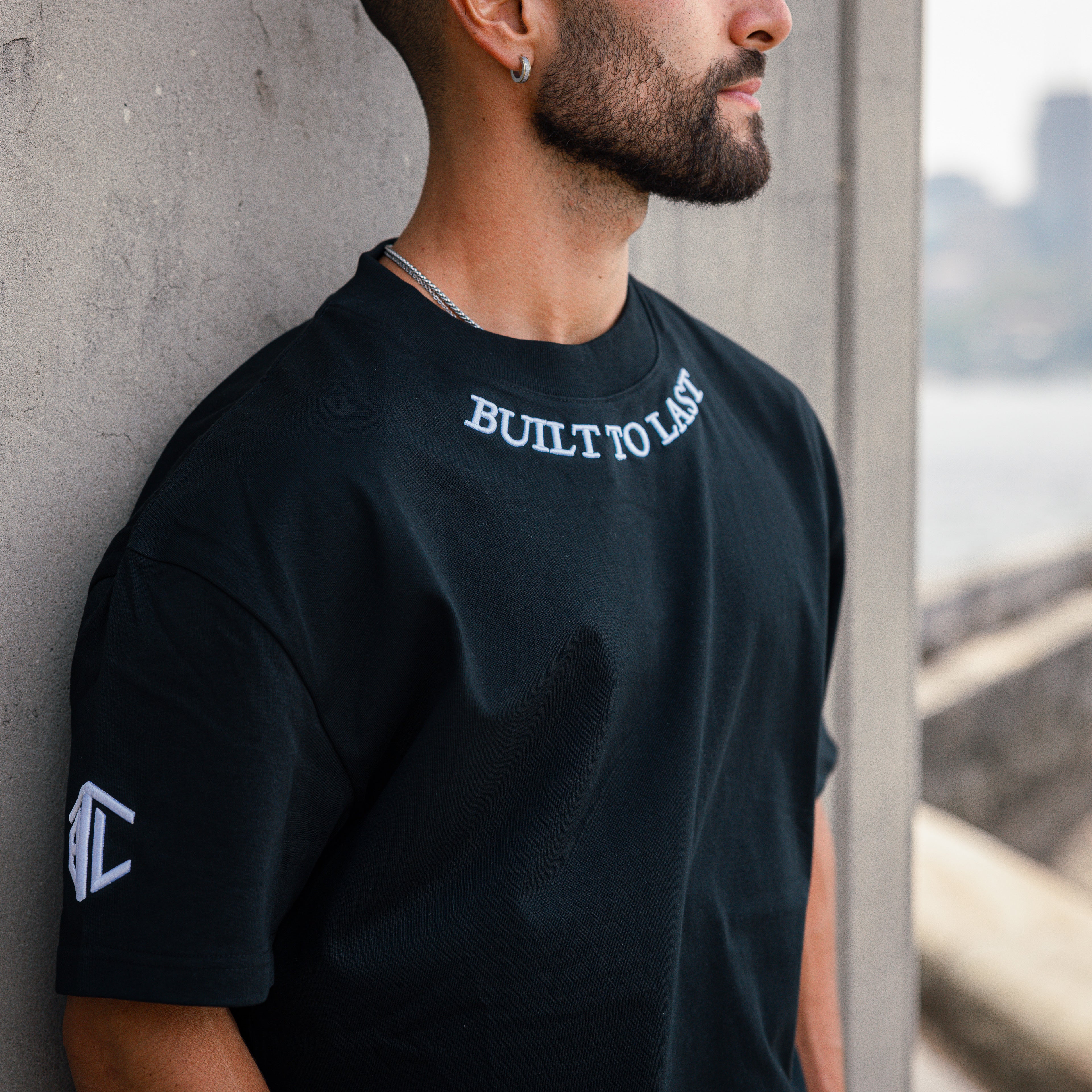 A man wearing a black crewneck t-shirt with 'BUILT TO LAST' printed in white on the chest and a white geometric logo on the sleeve, leaning against a concrete pillar.