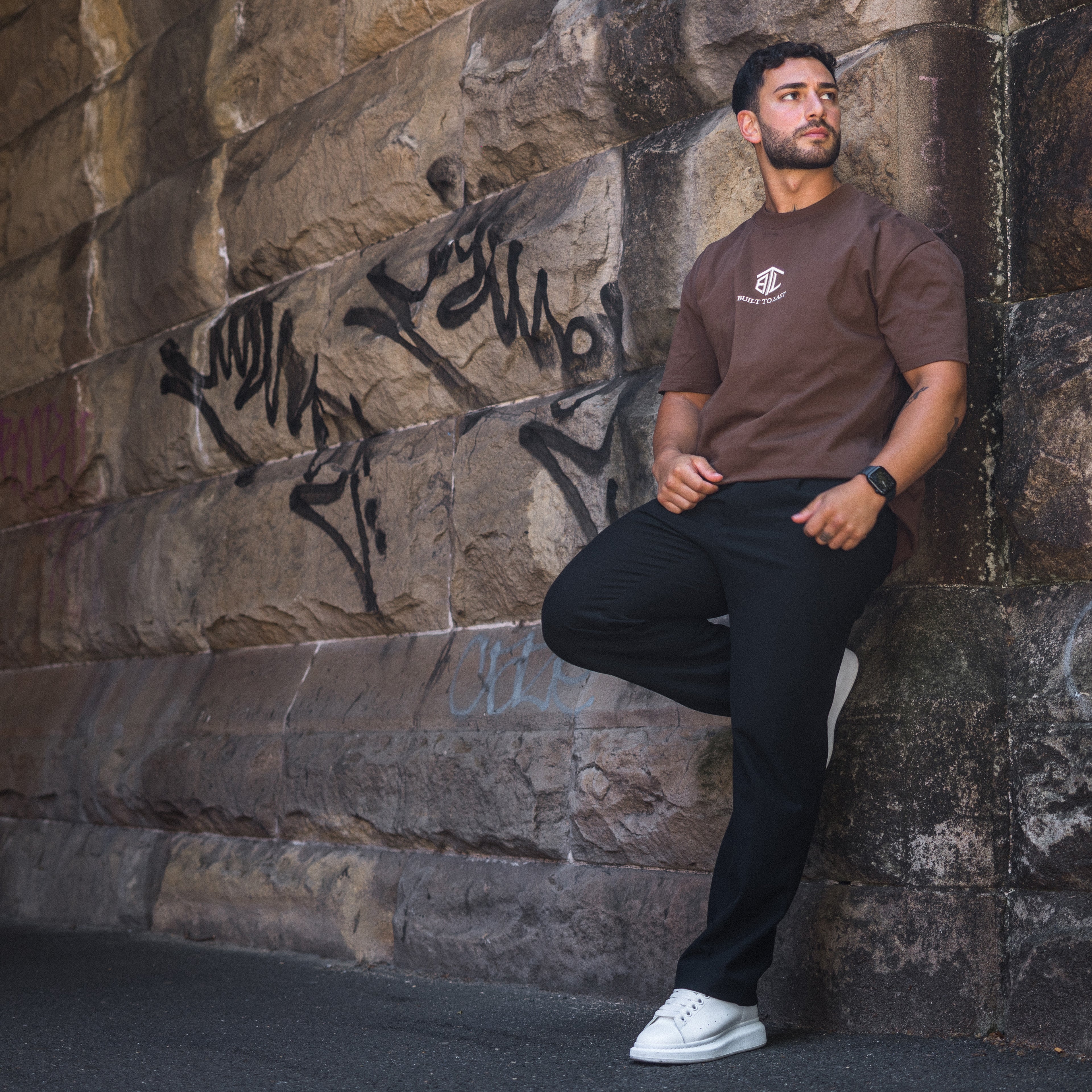Model leaning the wall wearing Built to Last Eternal Tee Chocolate heavyweight cotton T-shirt with embroidered chest logo