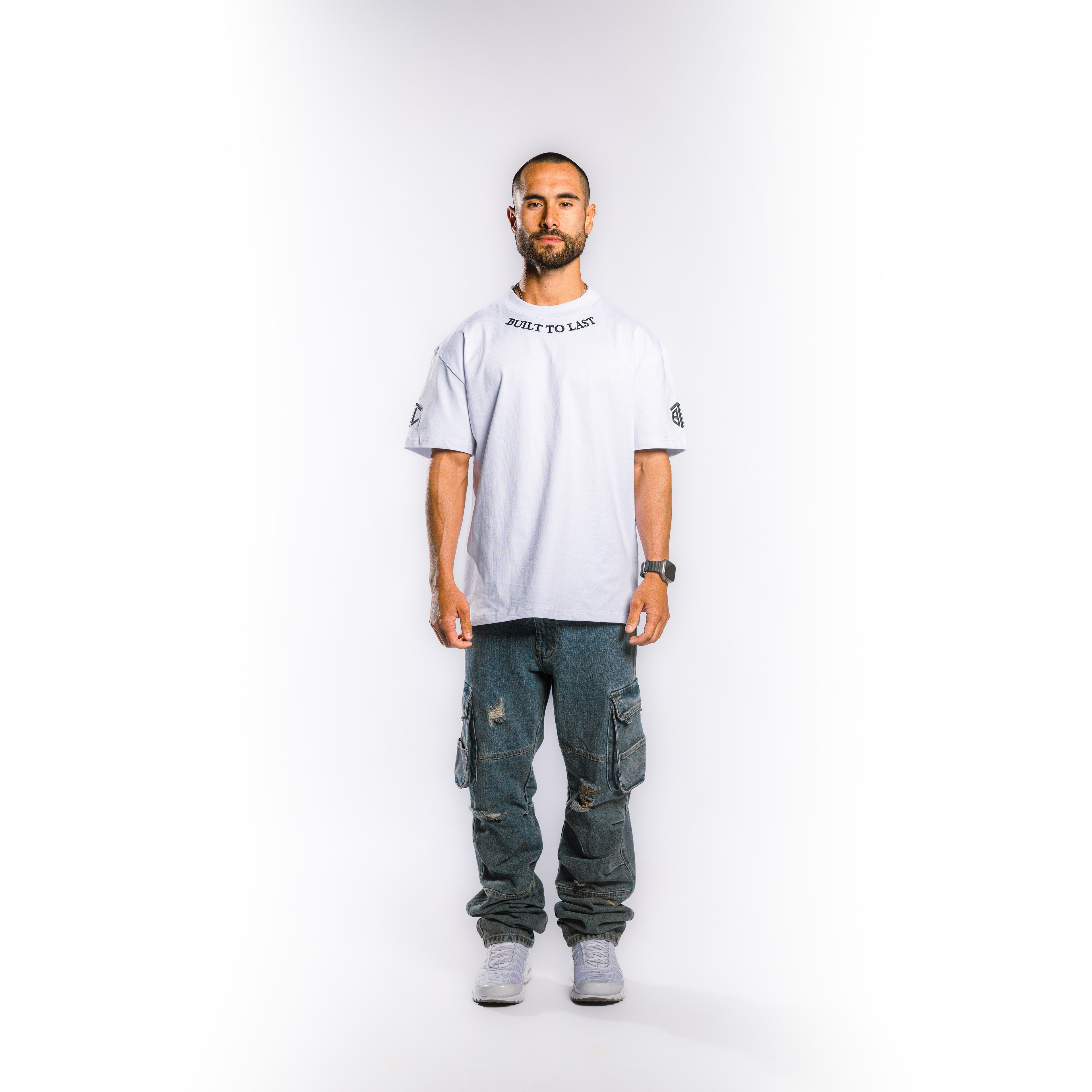 A man stands against a light gray studio background wearing a white oversized T-shirt with black text on the chest and sleeve patches, dark distressed cargo pants, and light gray sneakers.