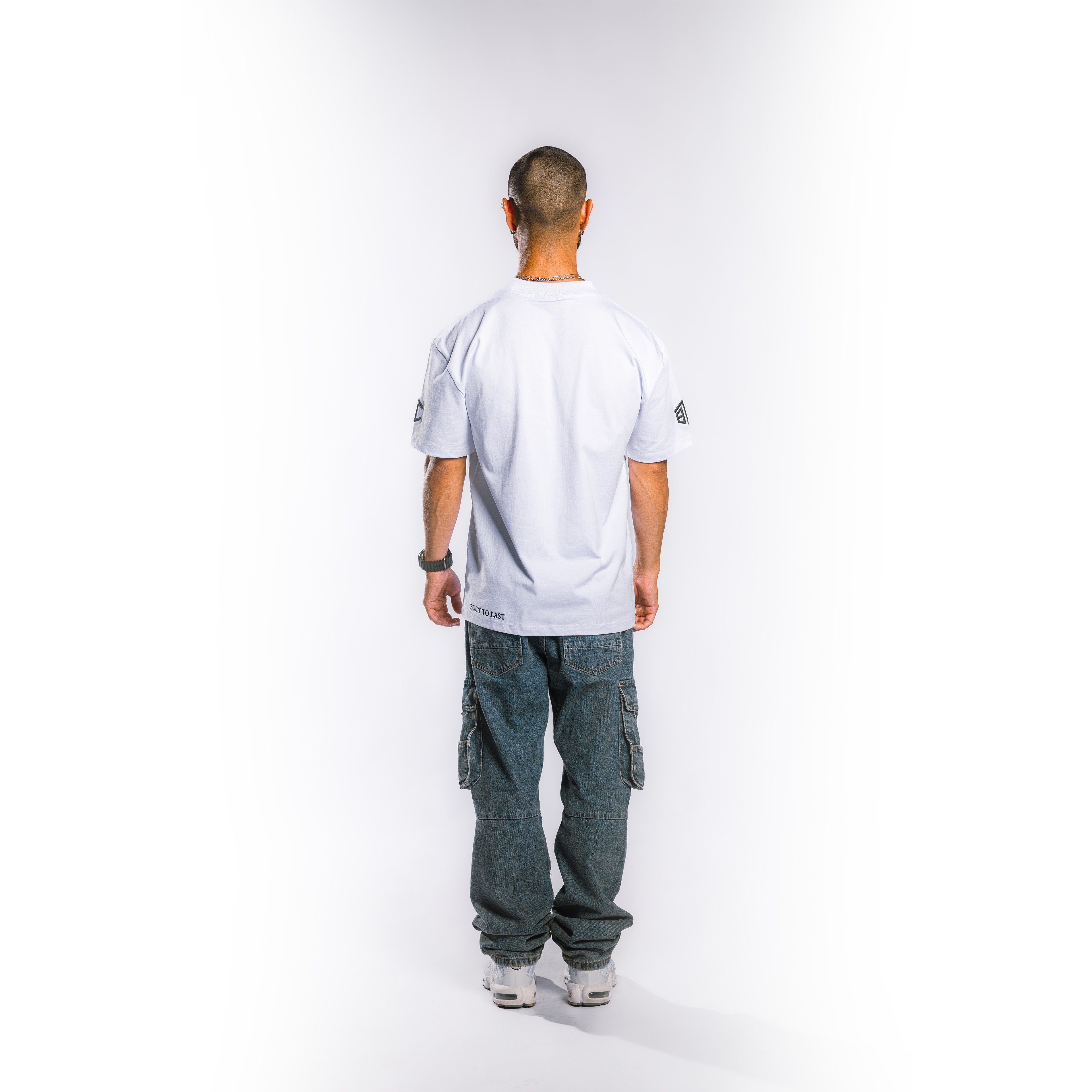 A man viewed from behind wearing a white short-sleeve athletic jersey, dark gray cargo pants, and white sneakers, standing against a plain white studio background.