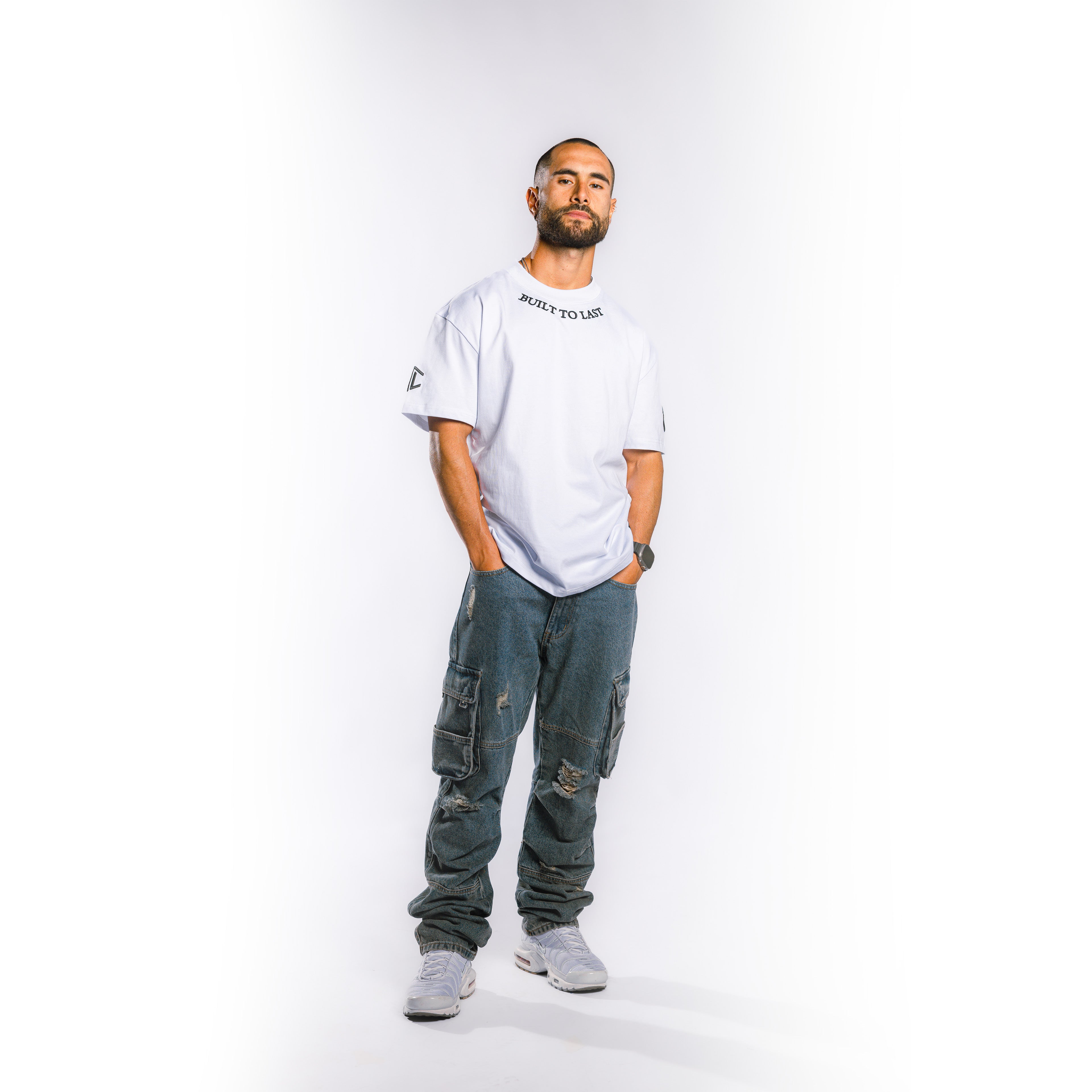 Model wearing a white Built To Last Clothing t-shirt with collar logo and distressed denim cargo pants against a white background.