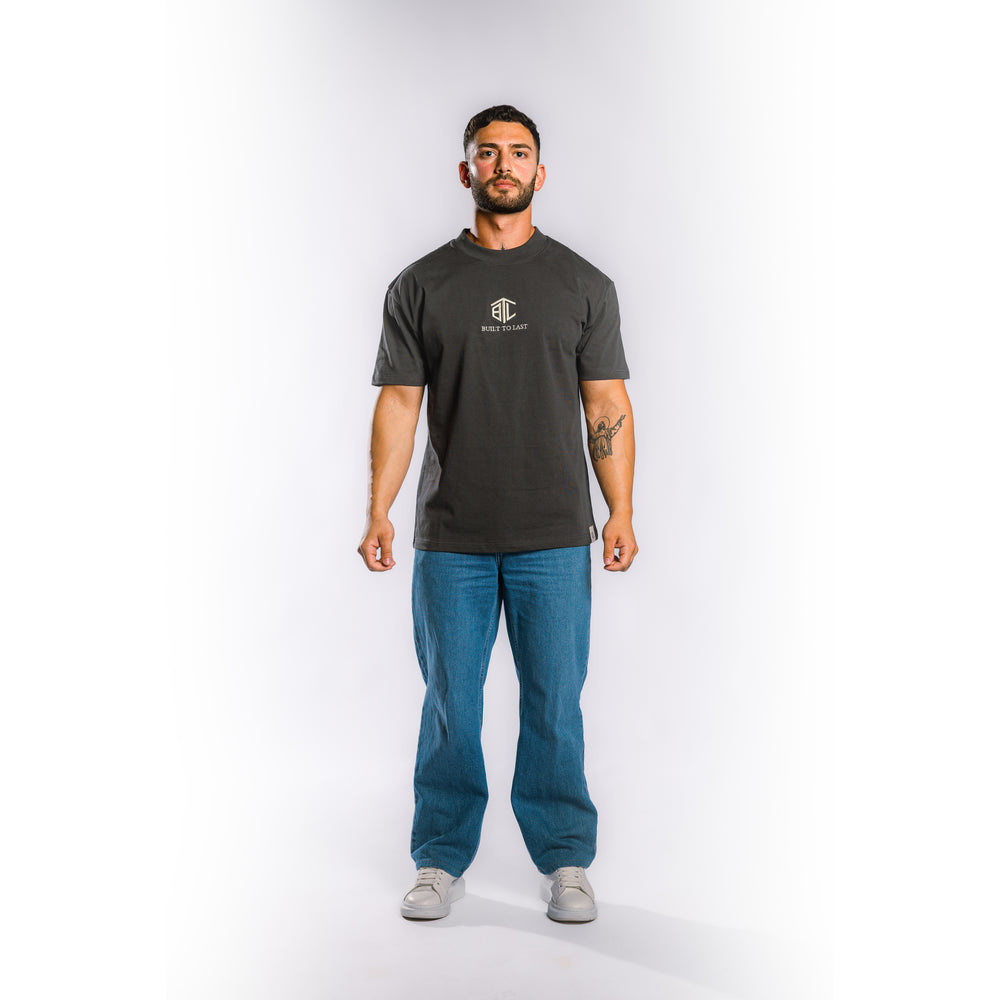 A man modeling a dark charcoal gray crewneck t-shirt with a small white chest logo, paired with blue jeans and gray sneakers, standing against a light gray studio background.