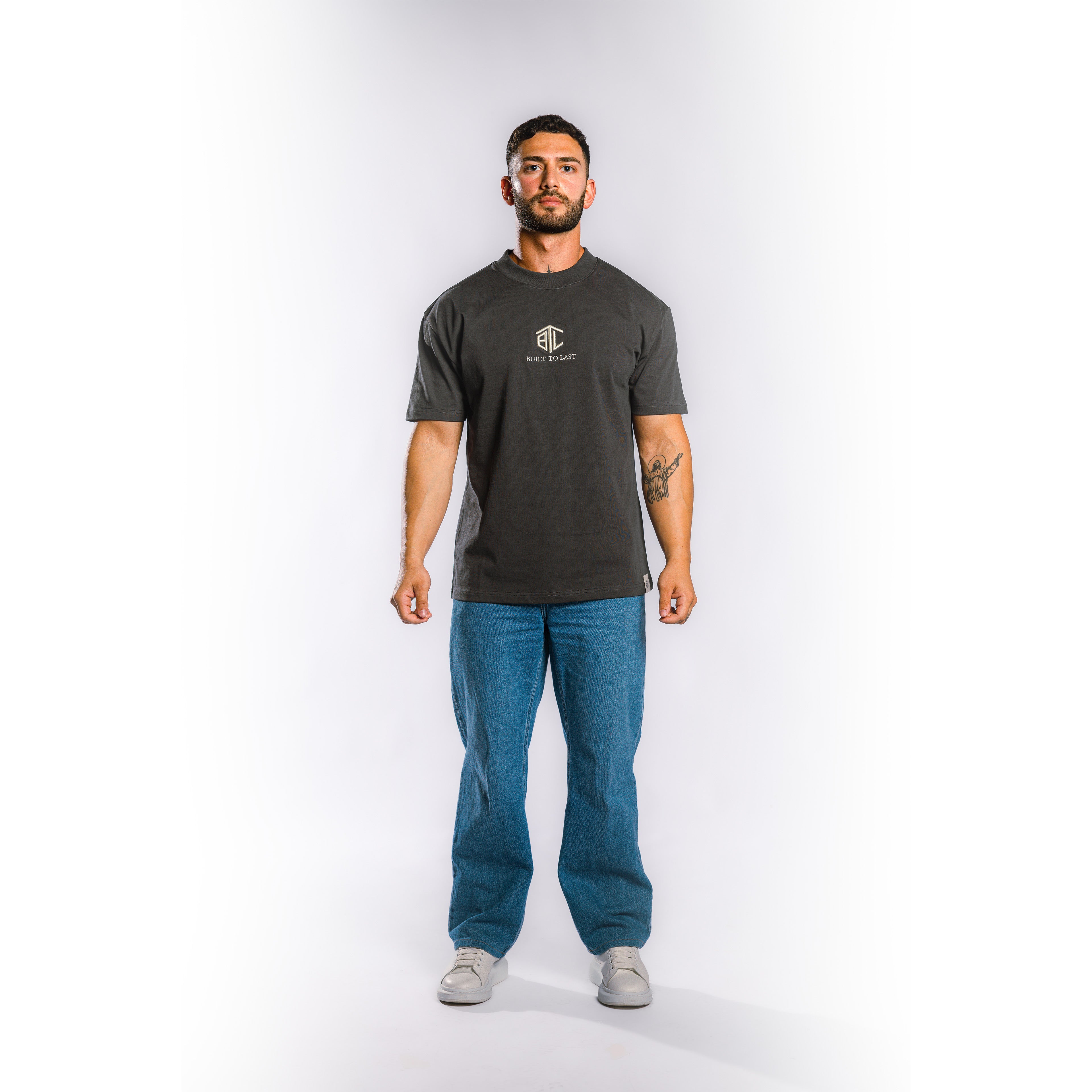 A man modeling a dark charcoal gray crewneck t-shirt with a small white chest logo, paired with blue jeans and gray sneakers, standing against a light gray studio background.